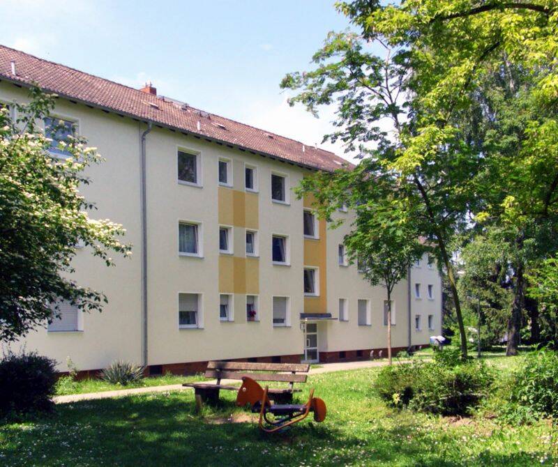 Hausansicht eines mehrstöckigen Wohngebäudes umgeben von Wiese, Sträuchern und Bäumen. 